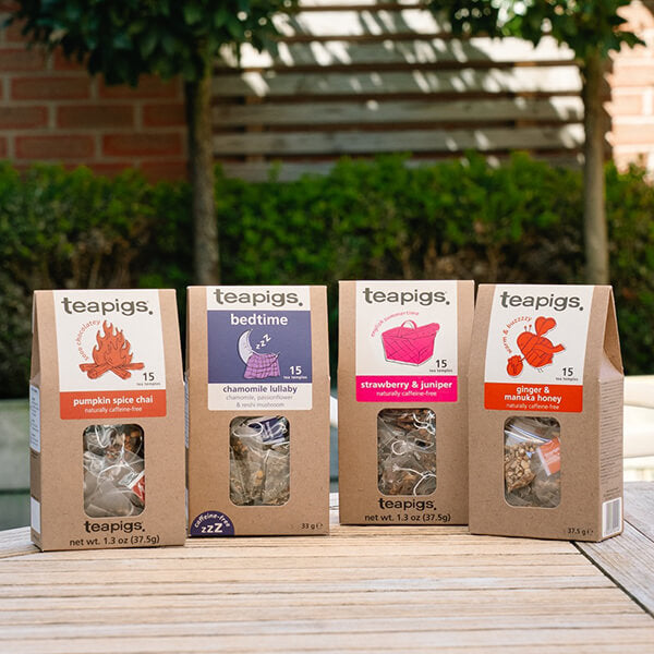 teapigs herbal and caffeine-free tea gift assortment featuring four boxed flavors—Pumpkin Spice Chai, Bedtime Chamomile Lullaby, Strawberry & Juniper, and Ginger & Manuka Honey—displayed on a wooden outdoor table with a garden background.